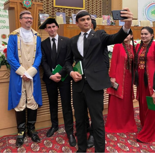 Greg Buford dressed in 18th century garb as program participants in traditional Turkmen dresses stand to the side for a selfie taken by a young man wearing a suit and Turkmen cap. 